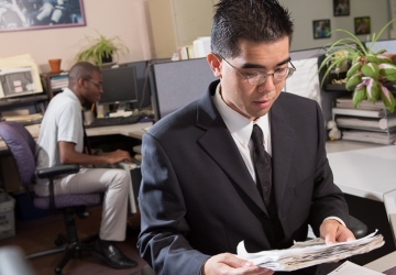 A young man in a suit works at a desk, while a co-worker files things behind him