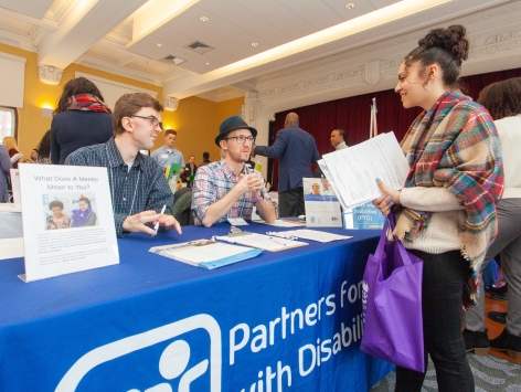 Two men sitting behind a table that has flyers on it, talking to a college-aged woman standing by the table.