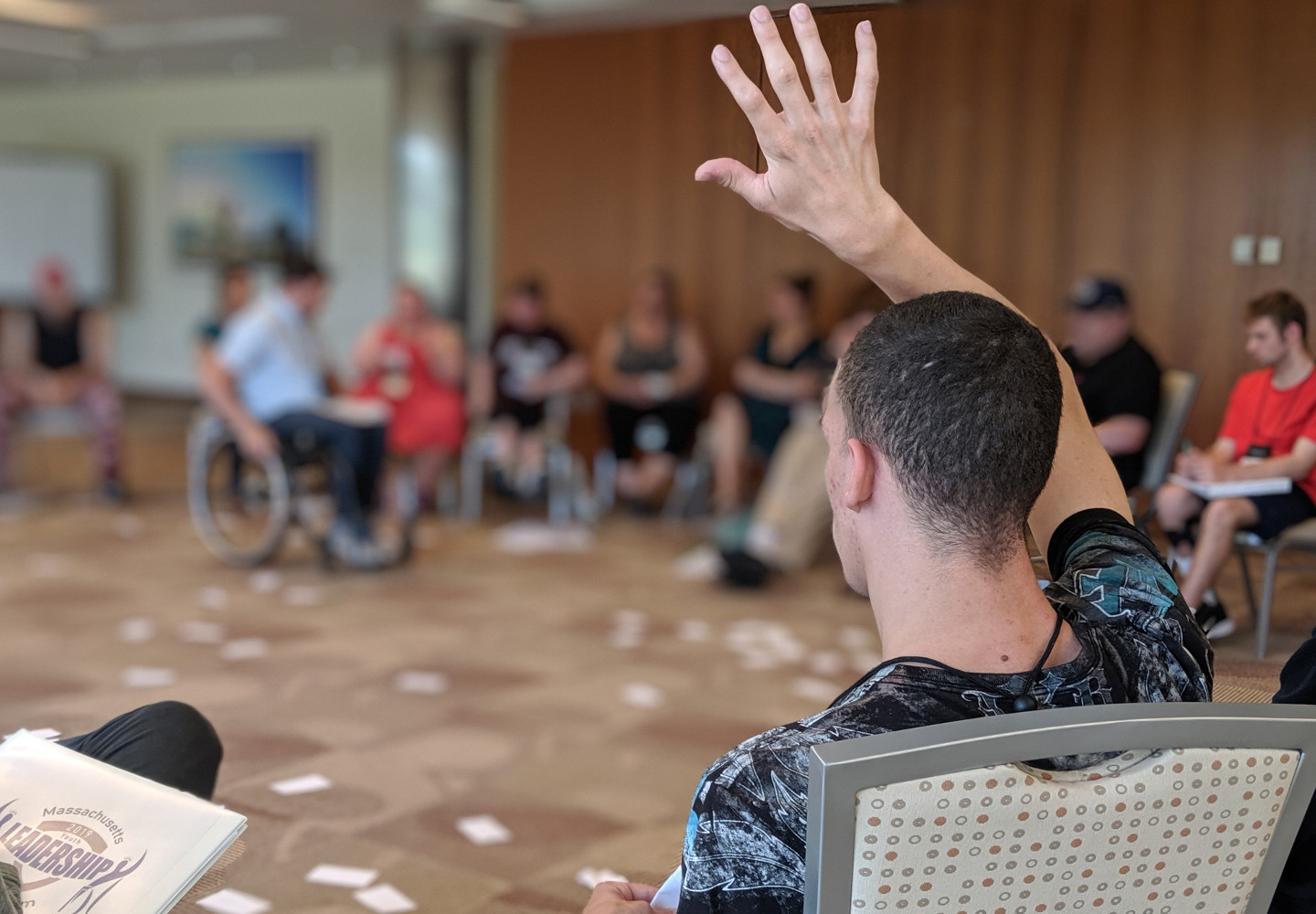 Young black man raises his hand to speak while sitting in a circle of his disabled peer