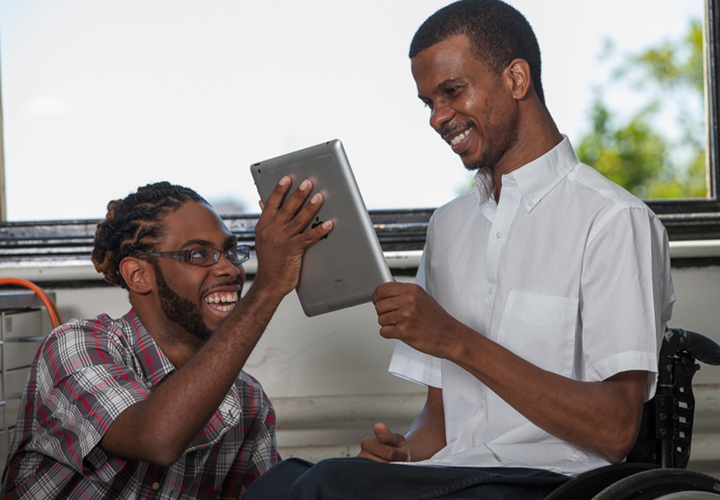 Young black man holds an iPad up for another young black man in a wheelchair