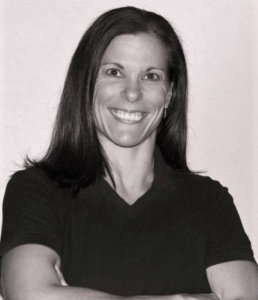 Black and white headshot of Amanda Kloo, a smiling woman with shoulder length dark hair, wearing a v-neck t-shirt.